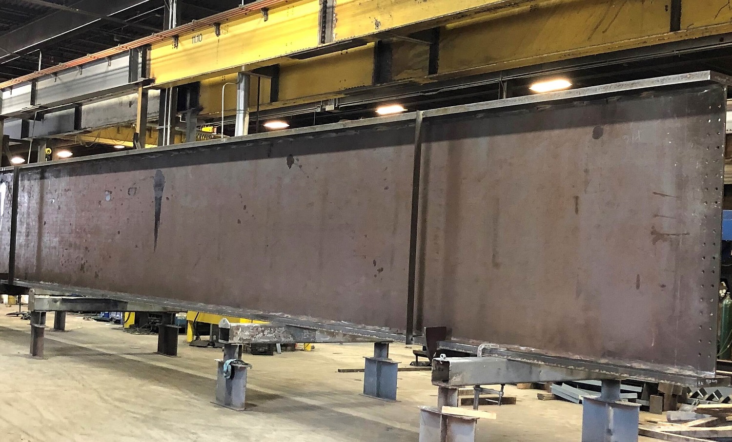 A long welded steel plate girder supported on stands inside a fabrication shop, with an overhead crane runway above it.