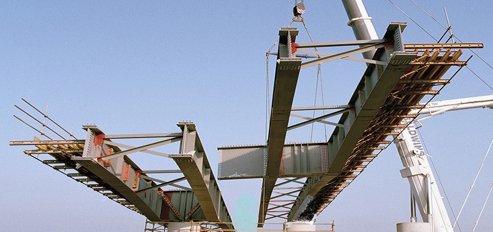 Steel plate girders and cross-bracing for a bridge span sit on a pier with deck formwork extending both ways, a crane hook lowering above them against a clear blue sky.