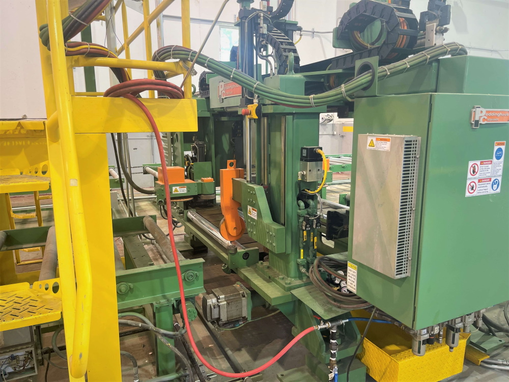 A green Peddinghaus CNC beam line with drilling head, control cabinet, red hydraulic hose and cable carrier chain, framed by yellow handrails.