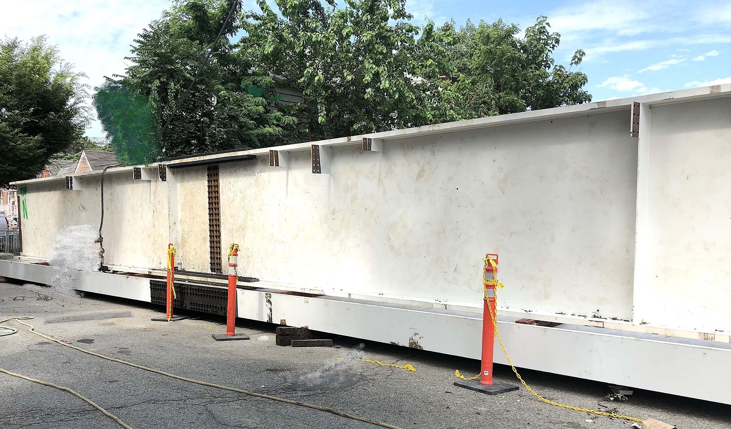 A tall white primed steel plate girder with stiffener plates staged on blocking at a kerbside, marked off with orange posts.