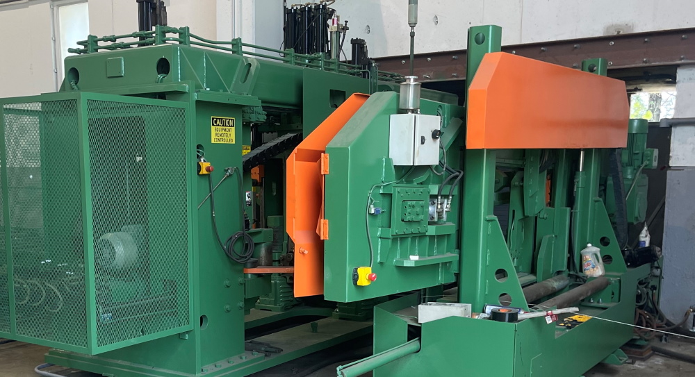 A wide view of a green and orange beam saw and drill line with mesh guard cabinets and an infeed table running the width of the frame.