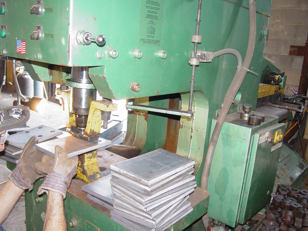 Gloved hands feed a steel base plate into a green hydraulic punch, with a stack of finished punched plates resting beside the machine.