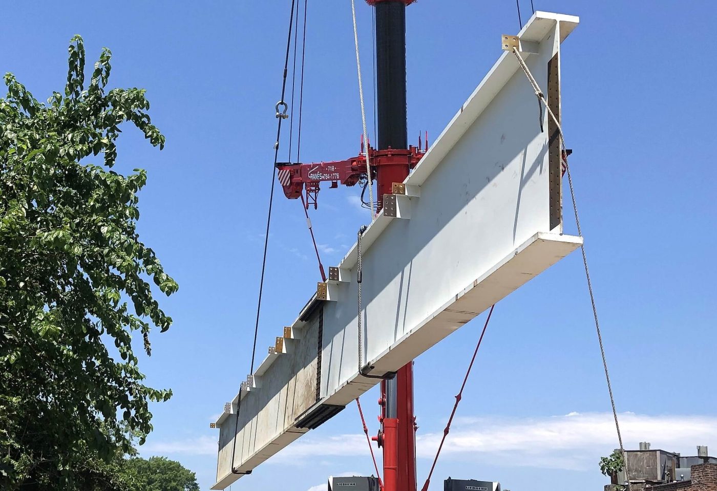 A white primed steel bridge girder hangs level from the wire ropes of a red mobile crane against a blue sky.