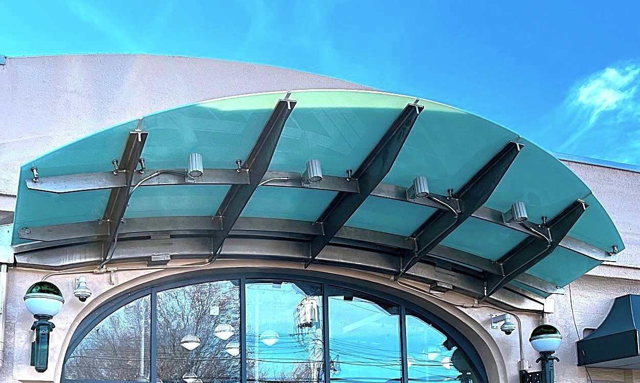 A curved green glass canopy carried on a row of tapered steel outrigger arms bolted to a building face above a tall arched storefront window.