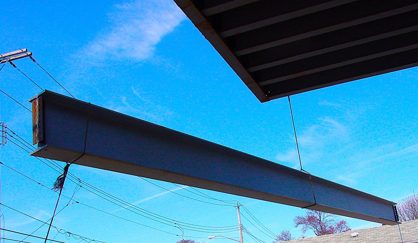 A long wide-flange steel beam bolted at one end and running out under a metal deck against a bright blue sky.
