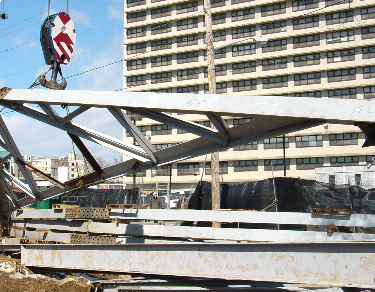 A long steel truss hangs from a crane hook above stacked steel members on a city job site, with a tall building behind.