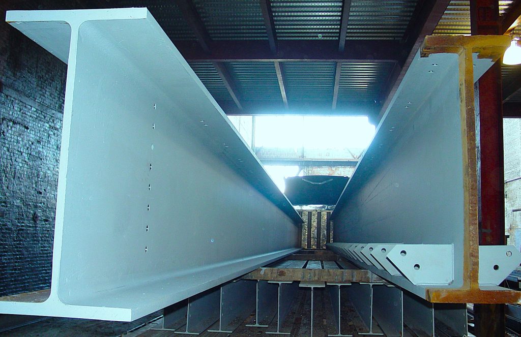 Two deep primed plate girders run the length of a fabrication shop, with stacked gusset plates alongside.