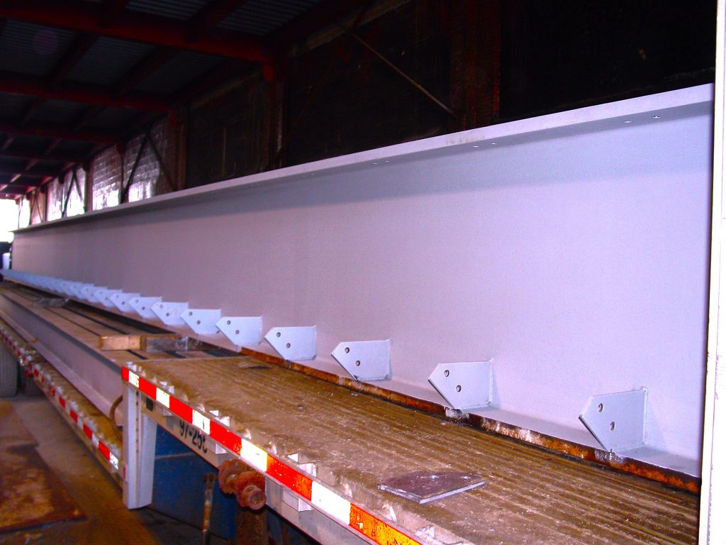 A long primed steel girder with welded clip angles sits on a flatbed trailer inside a fabrication shop.