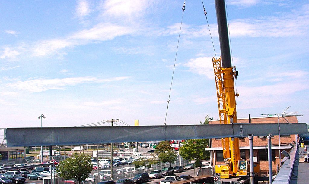 A long steel plate girder hangs level from the cables of a yellow truck crane above a parking lot.