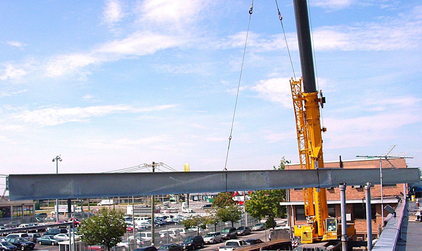 A long steel plate girder hangs level from the cables of a yellow truck crane above a parking lot.