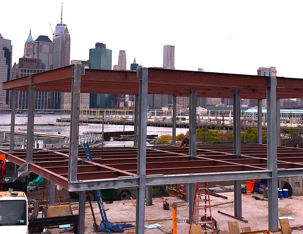A two-level steel frame with galvanized columns and red-oxide beams stands on a waterfront site with a city skyline across the water.