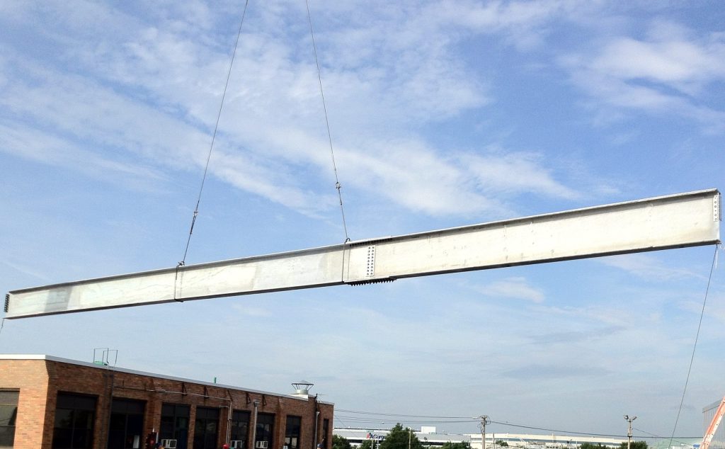 A galvanized steel girder with a bolted splice hangs from crane slings against a blue sky above a brick building.