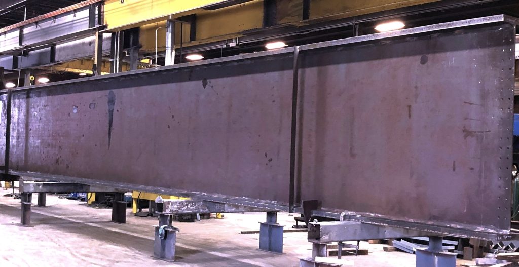 A deep built-up steel plate girder sits blocked up on short shop stands across a fabrication shop floor, with a yellow overhead crane runway and shop lights above it.
