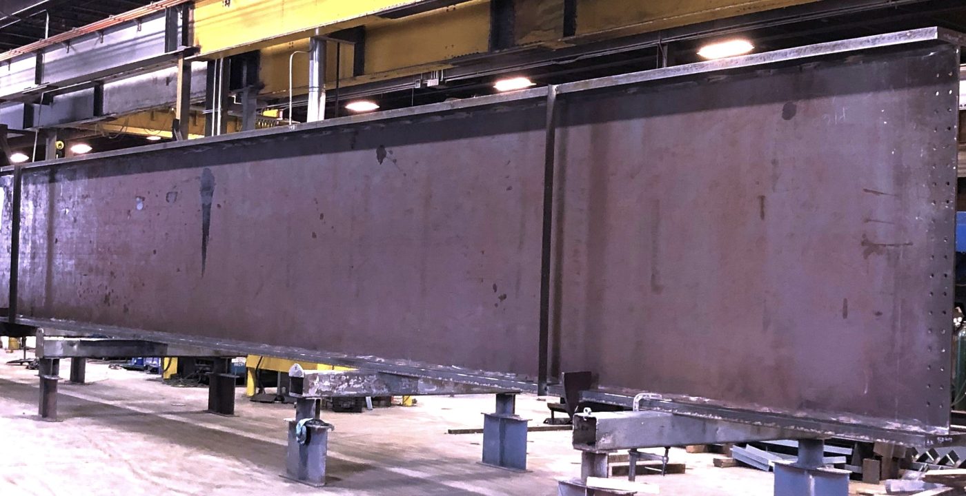 A deep built-up steel plate girder sits blocked up on short shop stands across a fabrication shop floor, with a yellow overhead crane runway and shop lights above it.