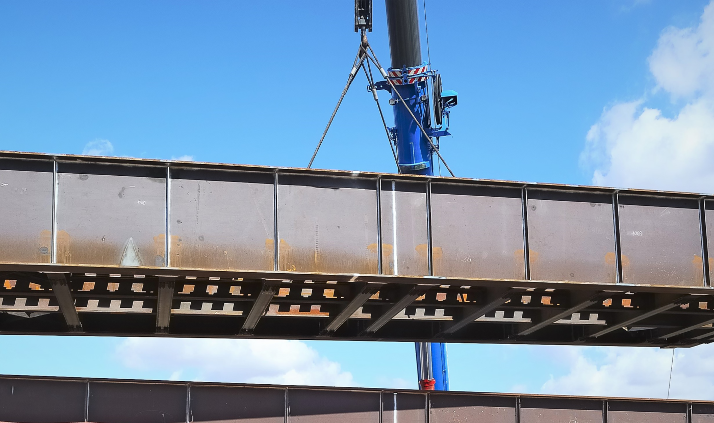 A steel bridge girder with rows of shear studs along its flange hanging from a blue mobile crane against a blue sky.