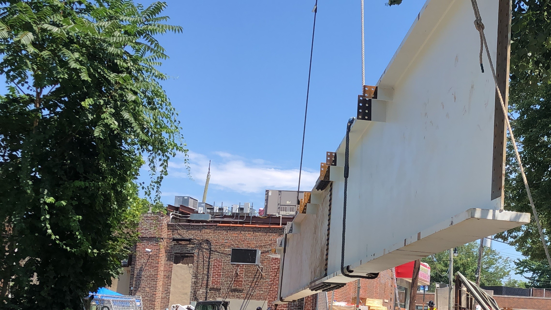 A white-primed steel plate girder hangs on wire rope slings above a brick building with rooftop HVAC units, with a tree at the left and a worker in a hard hat at the lower right of the street below.