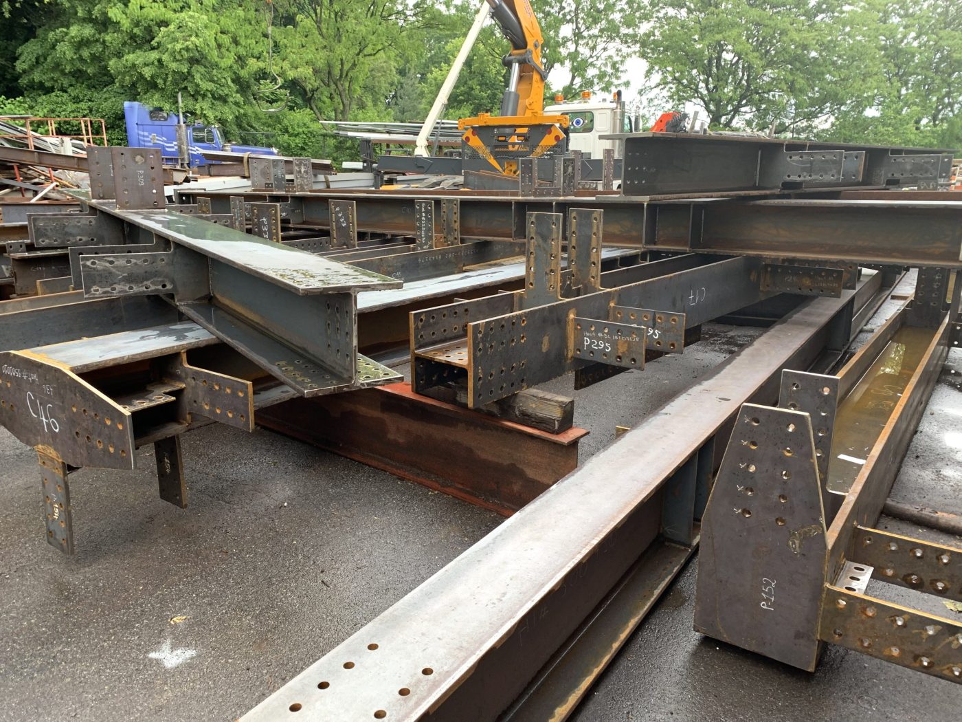 Rows of fabricated steel beams and columns with bolt holes and chalked piece marks laid out on a paved yard, with a boom truck loading steel behind them.