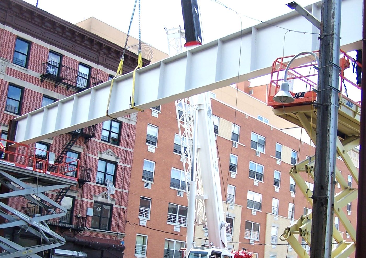 A painted steel girder hangs on slings between brick apartment buildings with boom lifts on either side.