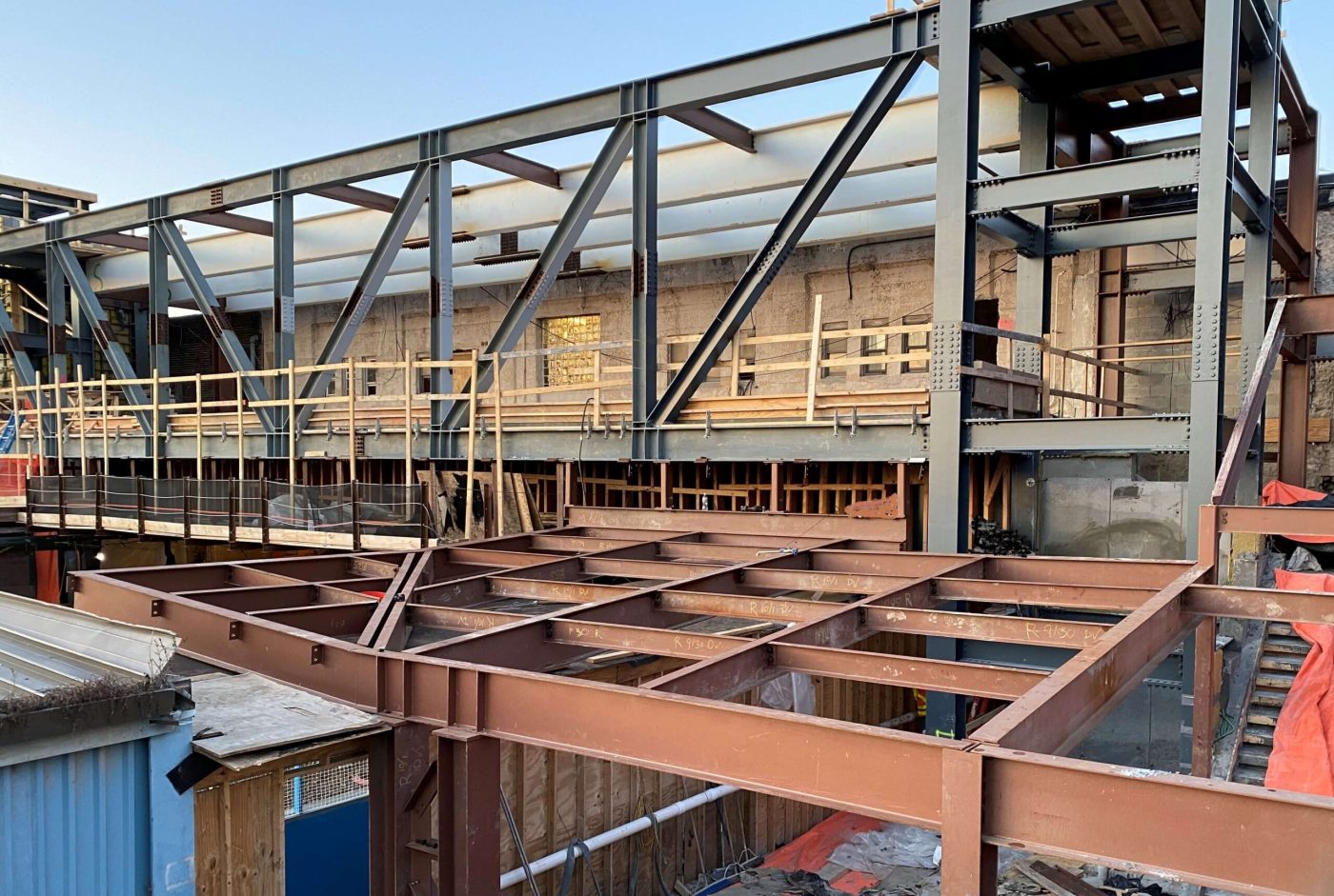 A structural steel building frame with bolted columns, diagonal bracing and red primed floor beams, with wood guardrails along the deck edge.