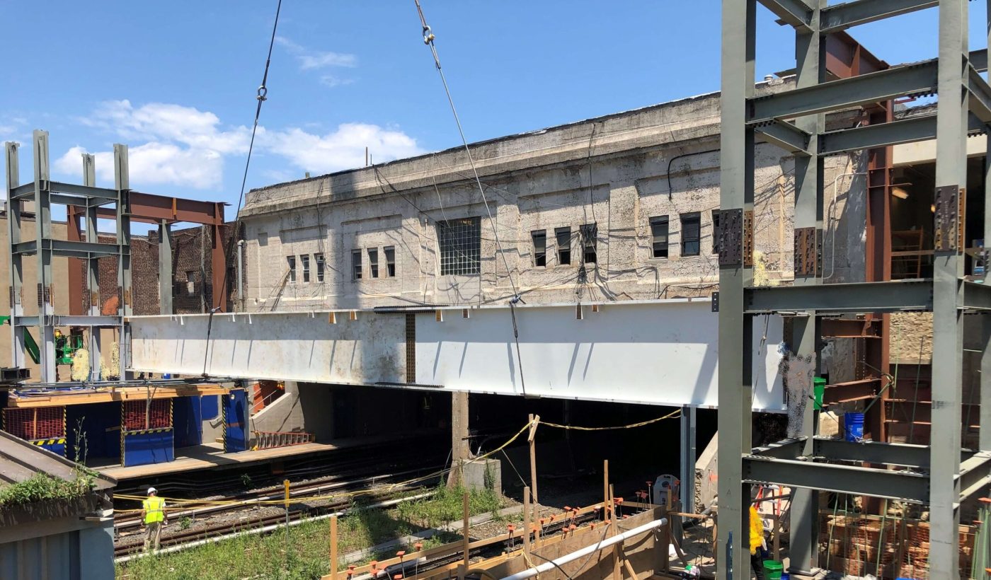 A long white painted plate girder hangs on crane lines above an open rail cut while a worker in a vest stands by the tracks below and a steel frame rises at the right.