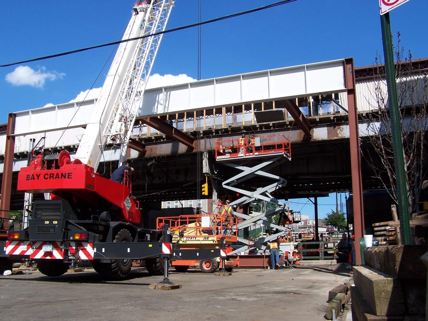 A red truck crane and two scissor lifts work beneath an elevated rail structure where new white painted girders have been set, with workers on the lift platforms.