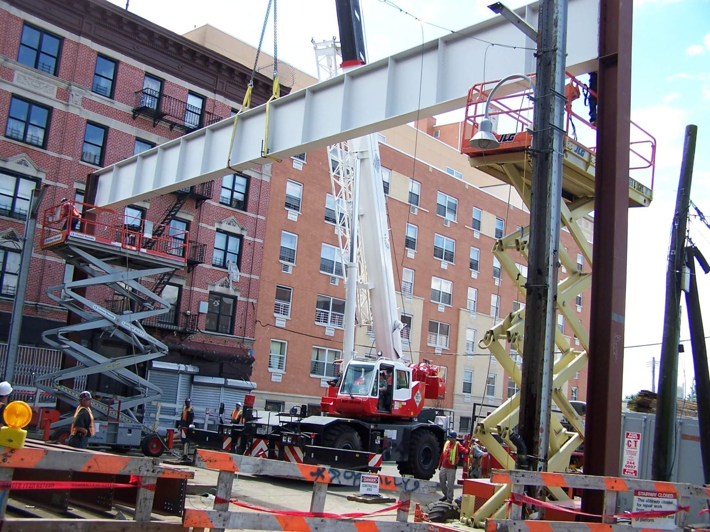 A rough terrain crane lifts a long white painted steel girder above a city street lined with brick buildings, with scissor lifts, barricades and ironworkers below.