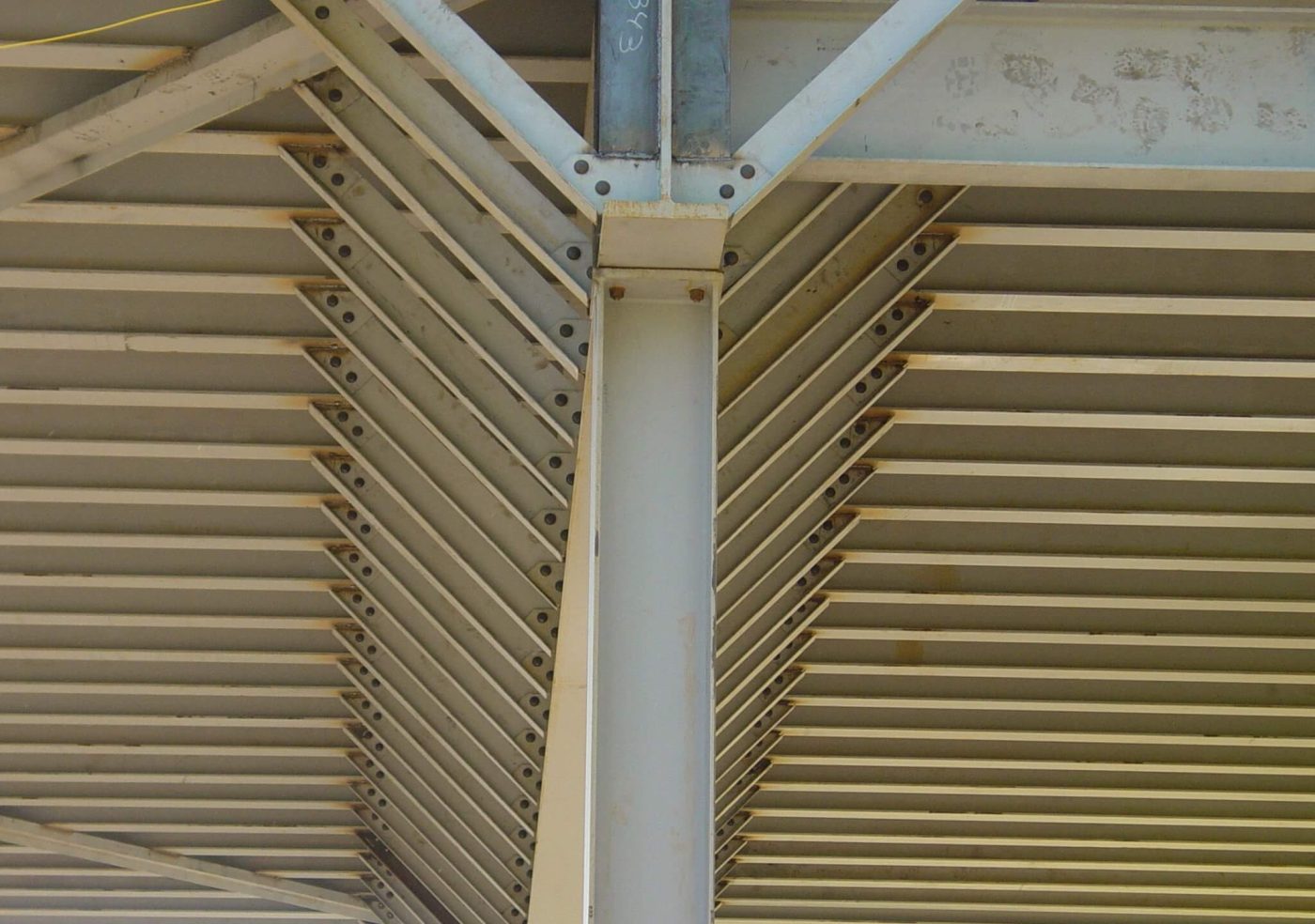 Looking up at the underside of a painted steel canopy, where bar joists fan out from a bolted beam to column connection to metal roof deck on both sides.