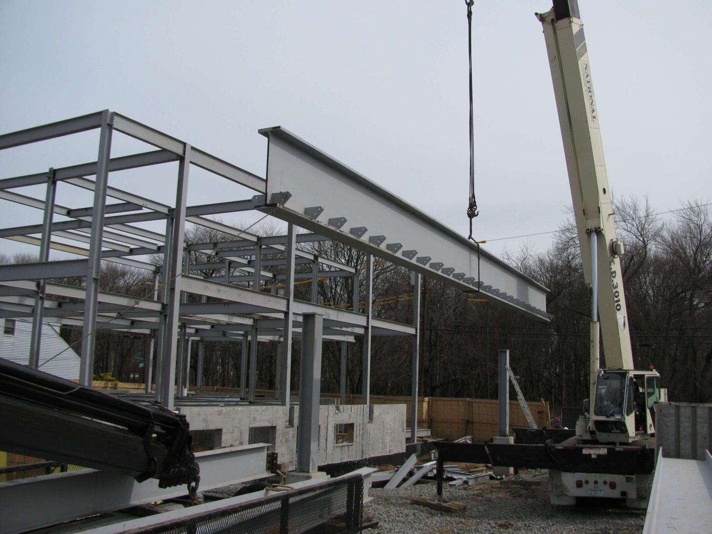 A truck-mounted hydraulic crane hoisting a long grey painted steel girder alongside a multi-bay steel building frame.