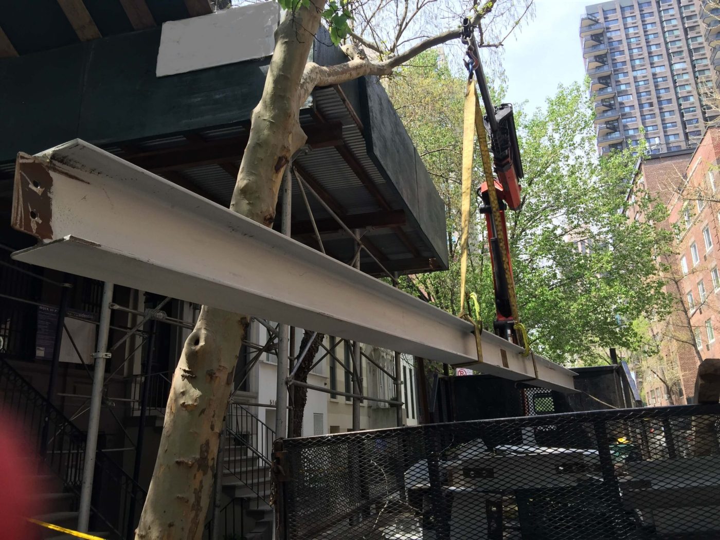 A long white-painted wide-flange steel beam held in yellow nylon slings by a red knuckle-boom crane on a narrow city street next to a scaffolded townhouse.