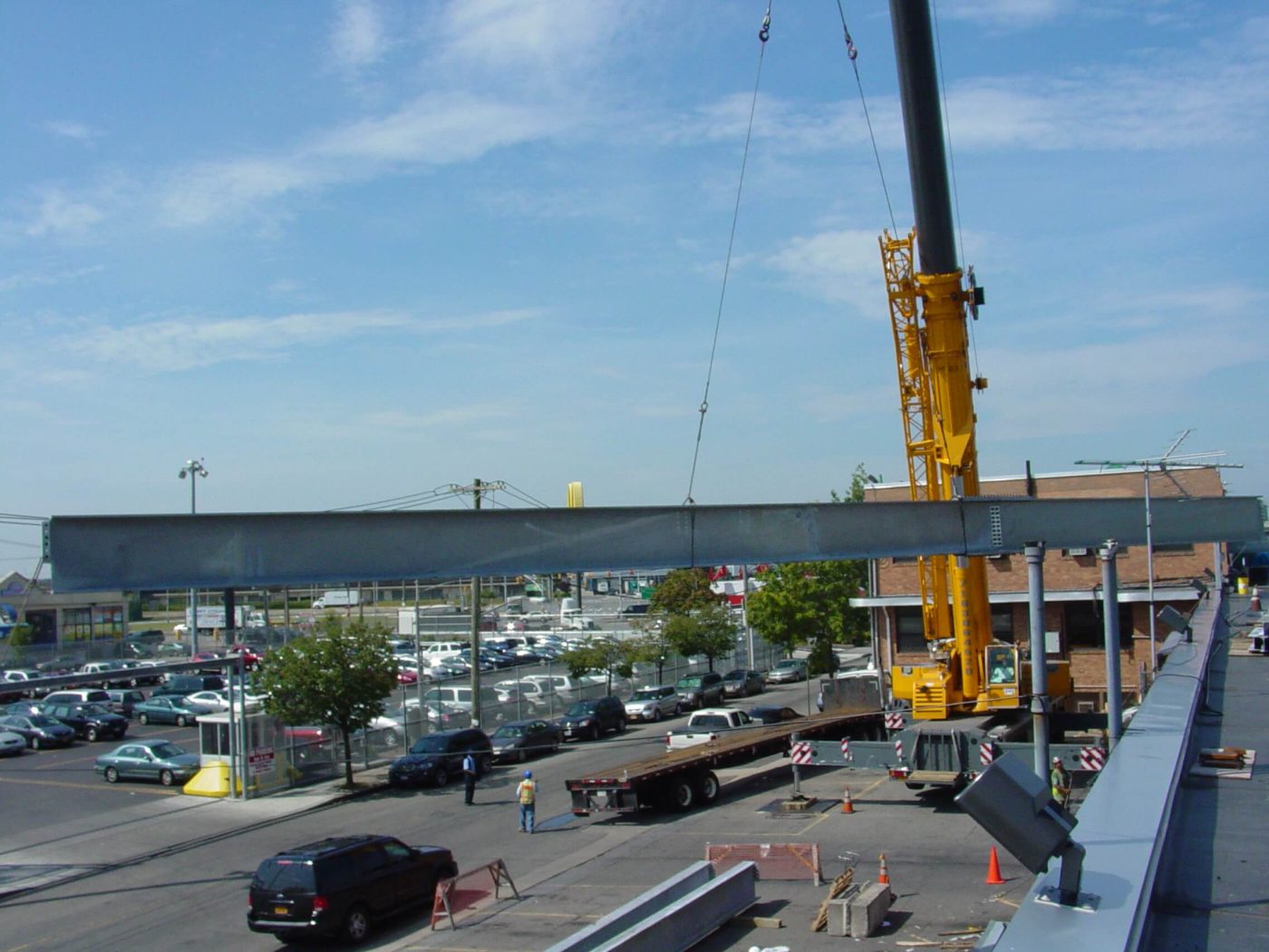 A yellow hydraulic crane lifting a long spliced steel girder high above a parking lot, with a flatbed trailer and two workers on the pavement below.