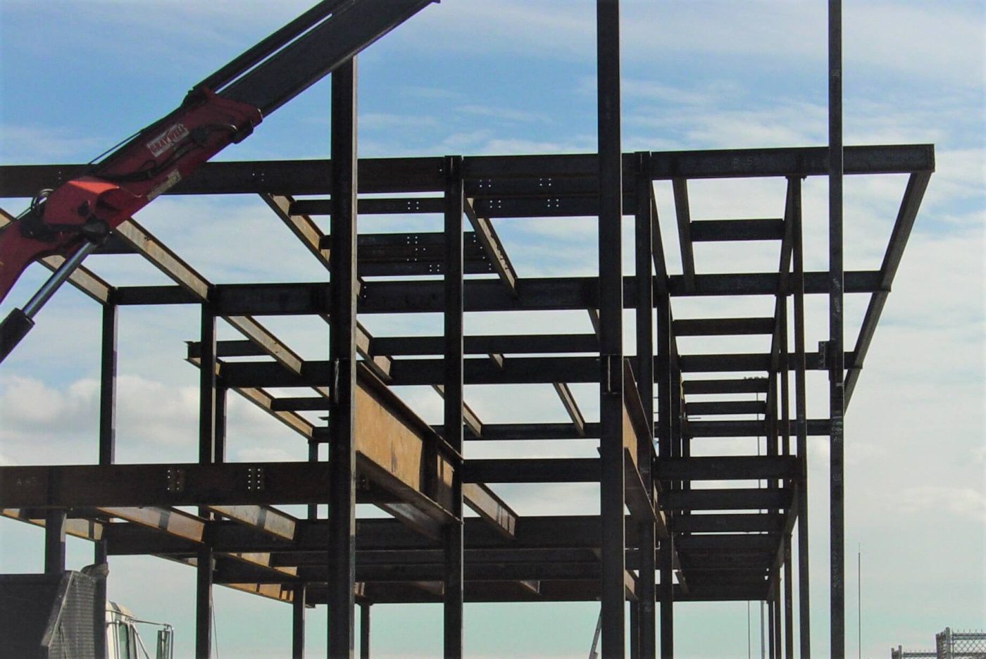 Steel stair stringers and landing framing inside a bolted structural steel frame, silhouetted against a pale blue sky with a crane boom at the left.