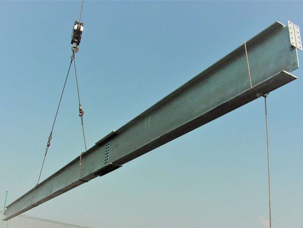 A long grey wide-flange steel beam with bolted splice plates, drilled end connection plates and a row of shear studs, hanging from a crane block against a clear blue sky.
