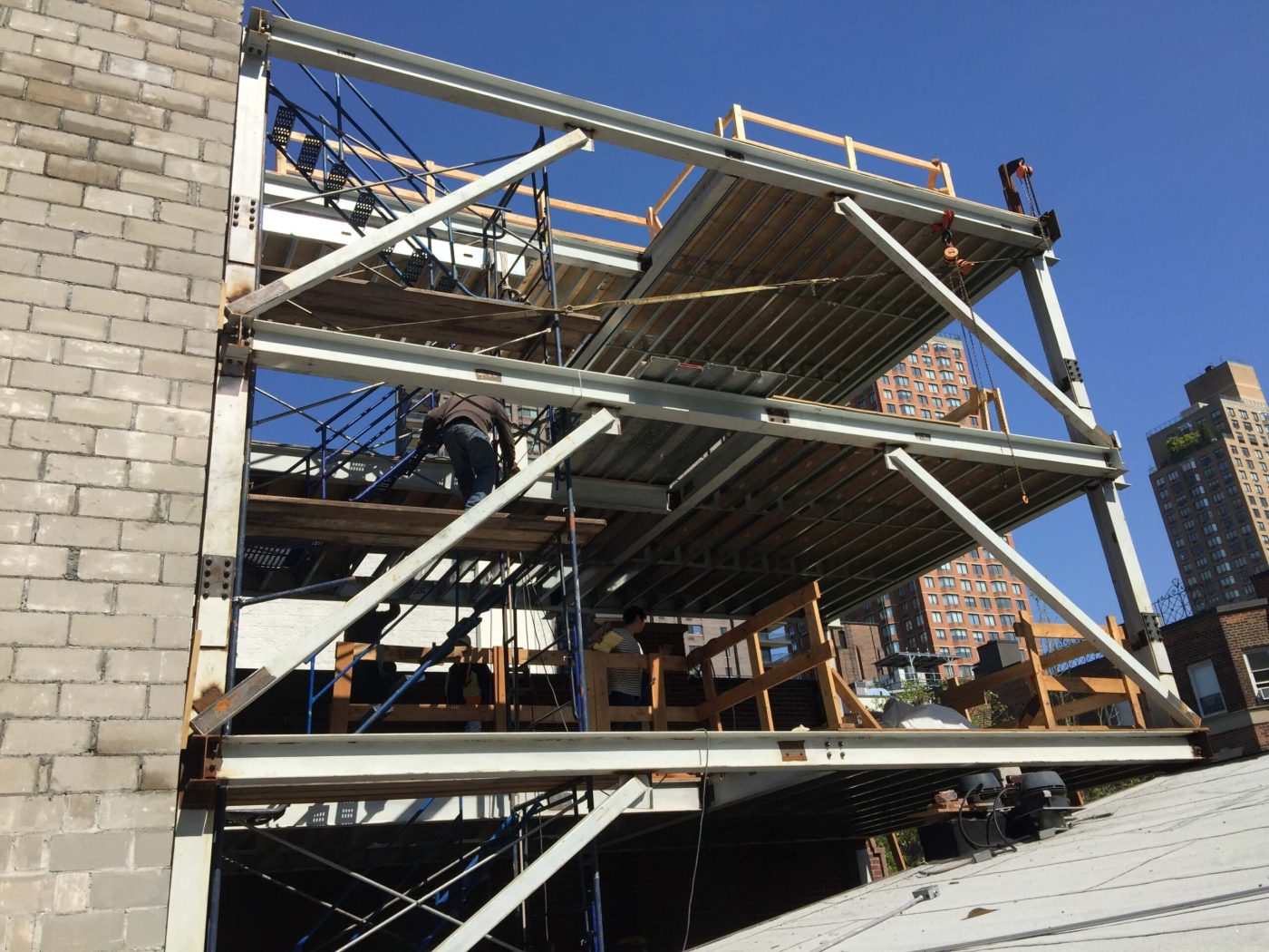 A multi-level steel extension frame built against a brick wall, with diagonal braces, metal floor deck, timber guardrails and ironworkers moving on the decks.