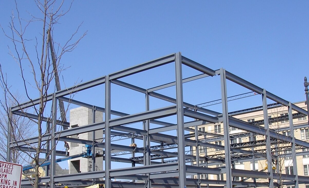 A grey painted steel building frame topped out against a clear blue sky, with bar joists filling the lower bays and a worker on a blue lift inside the structure.