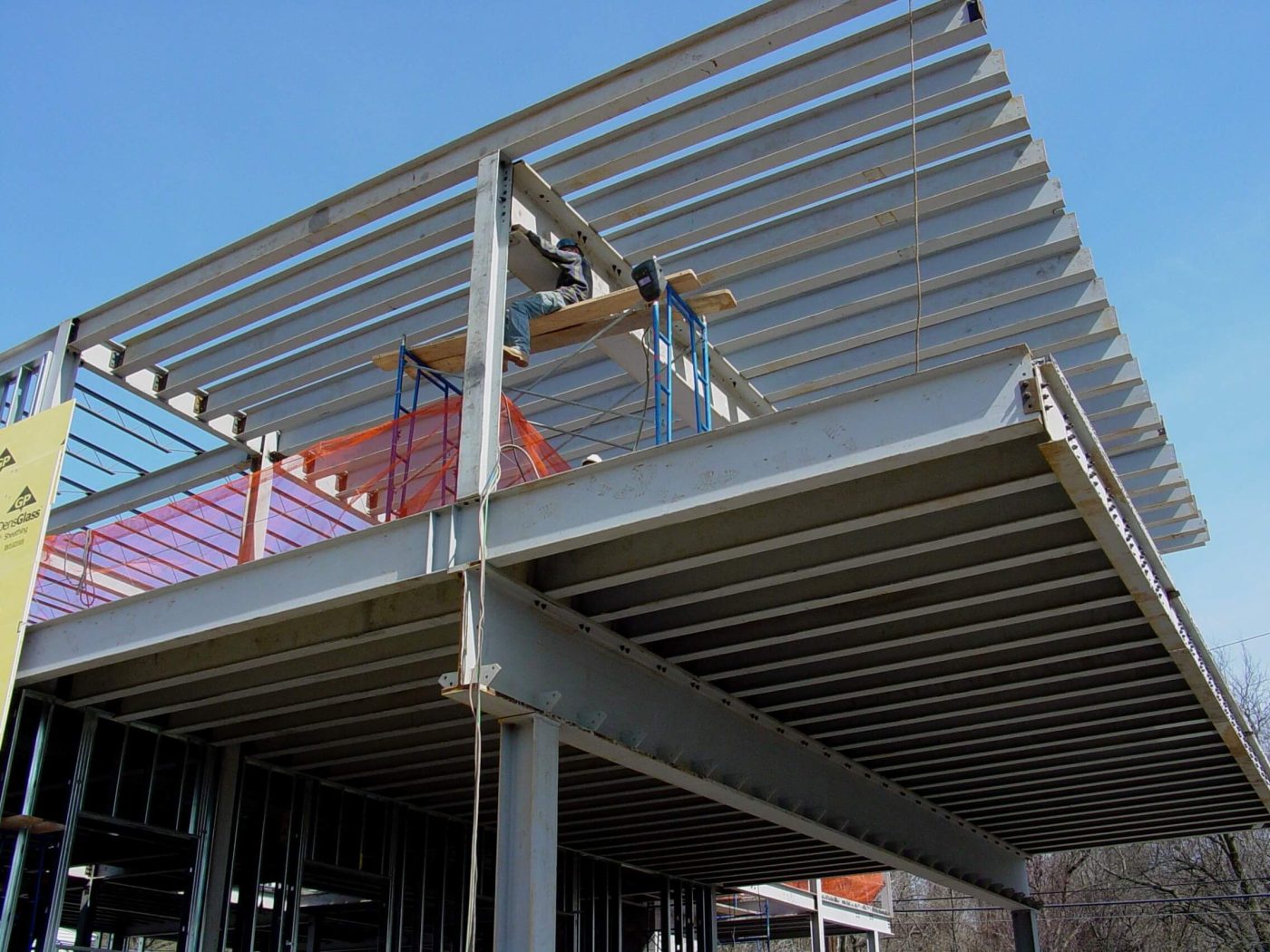 Cantilevered steel roof and floor framing with closely spaced infill beams against a blue sky, an ironworker in a hard hat working from a plank and scaffold at the beam line.