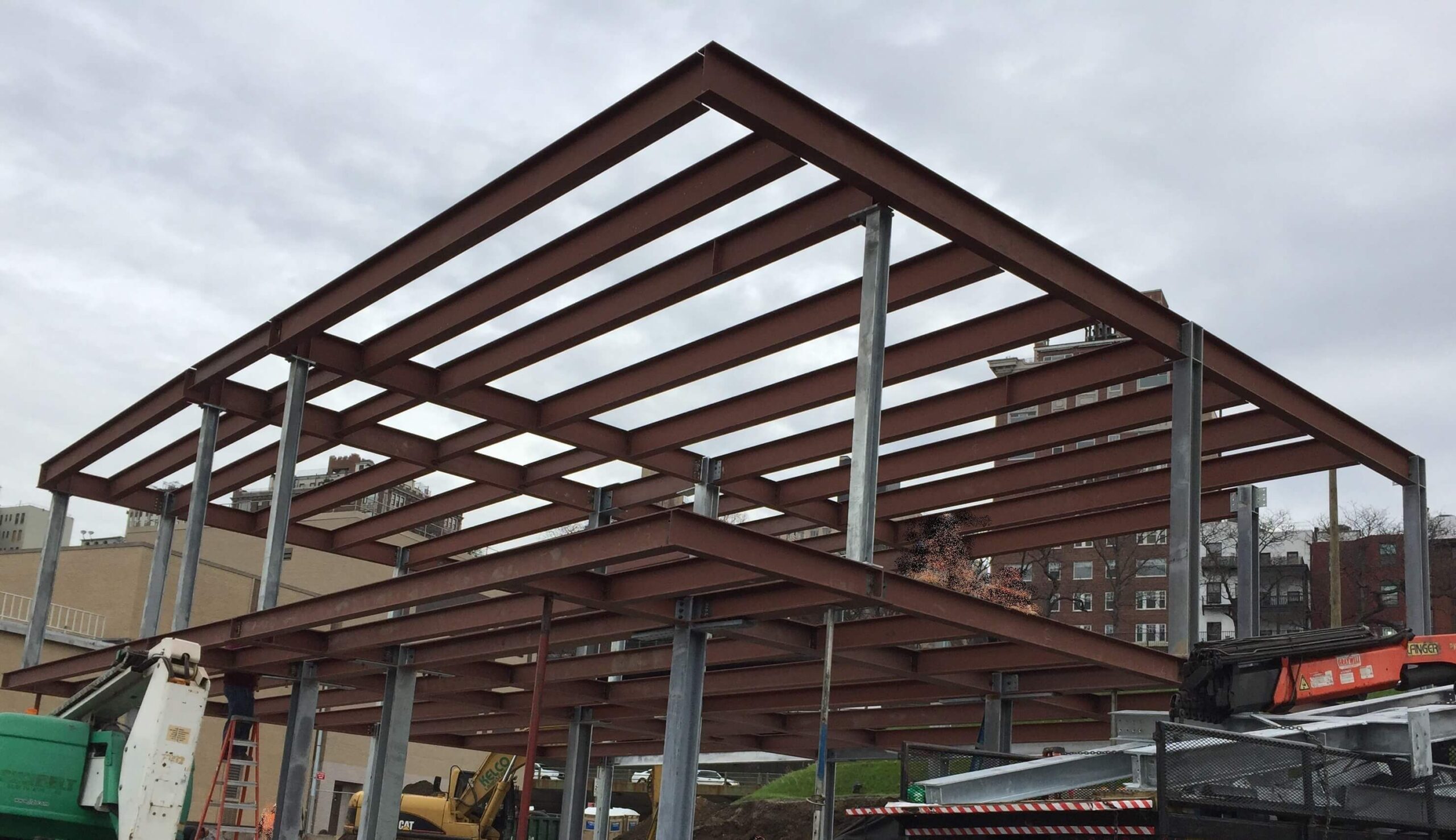 A pitched steel roof frame of red-primed rafters and a ridge beam supported on galvanized columns, with a boom lift and excavator working at ground level beneath.