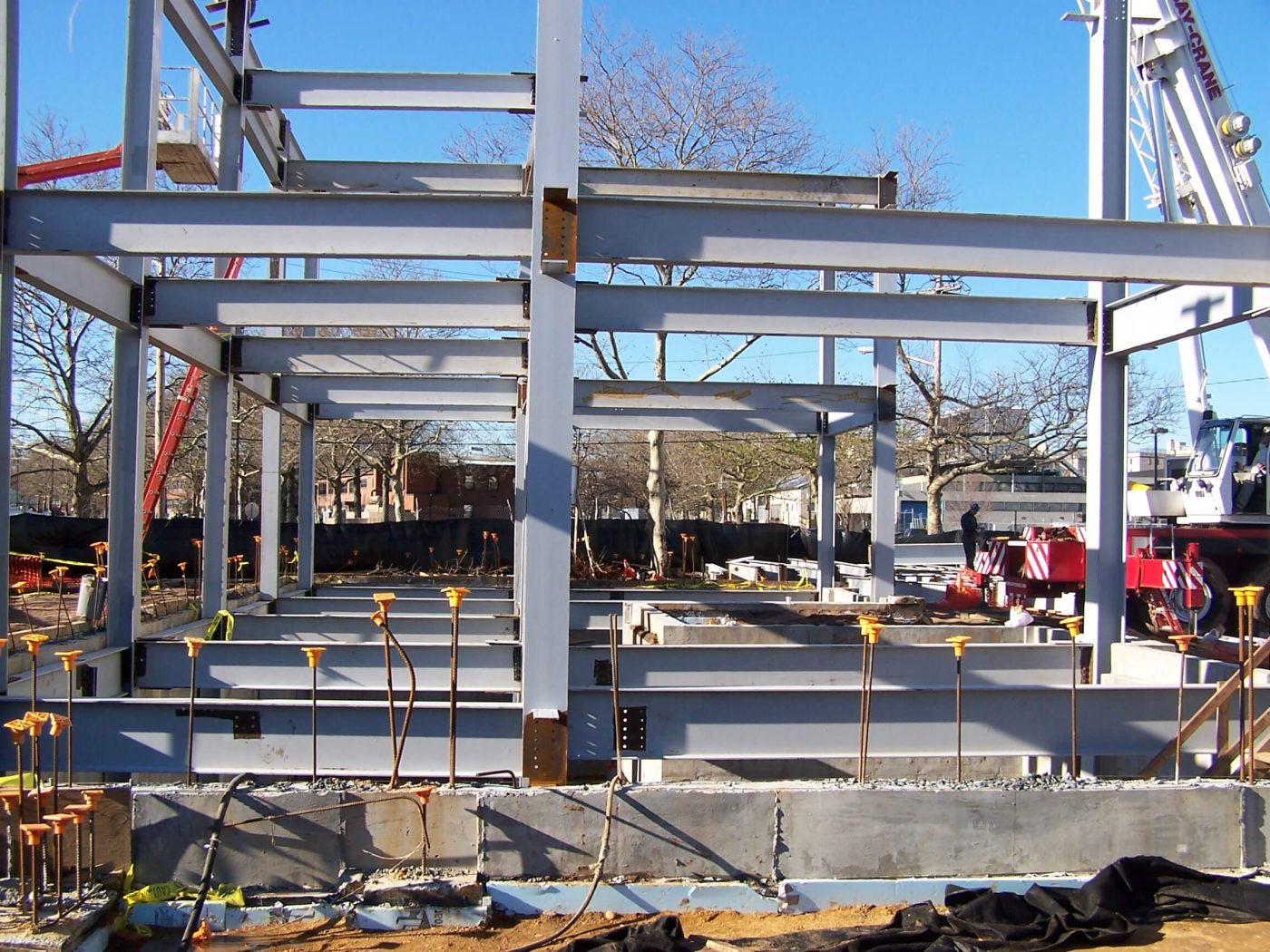 A bolted structural steel frame of columns and wide flange beams standing on concrete foundation walls under a clear blue sky, with a mobile crane parked at the right.