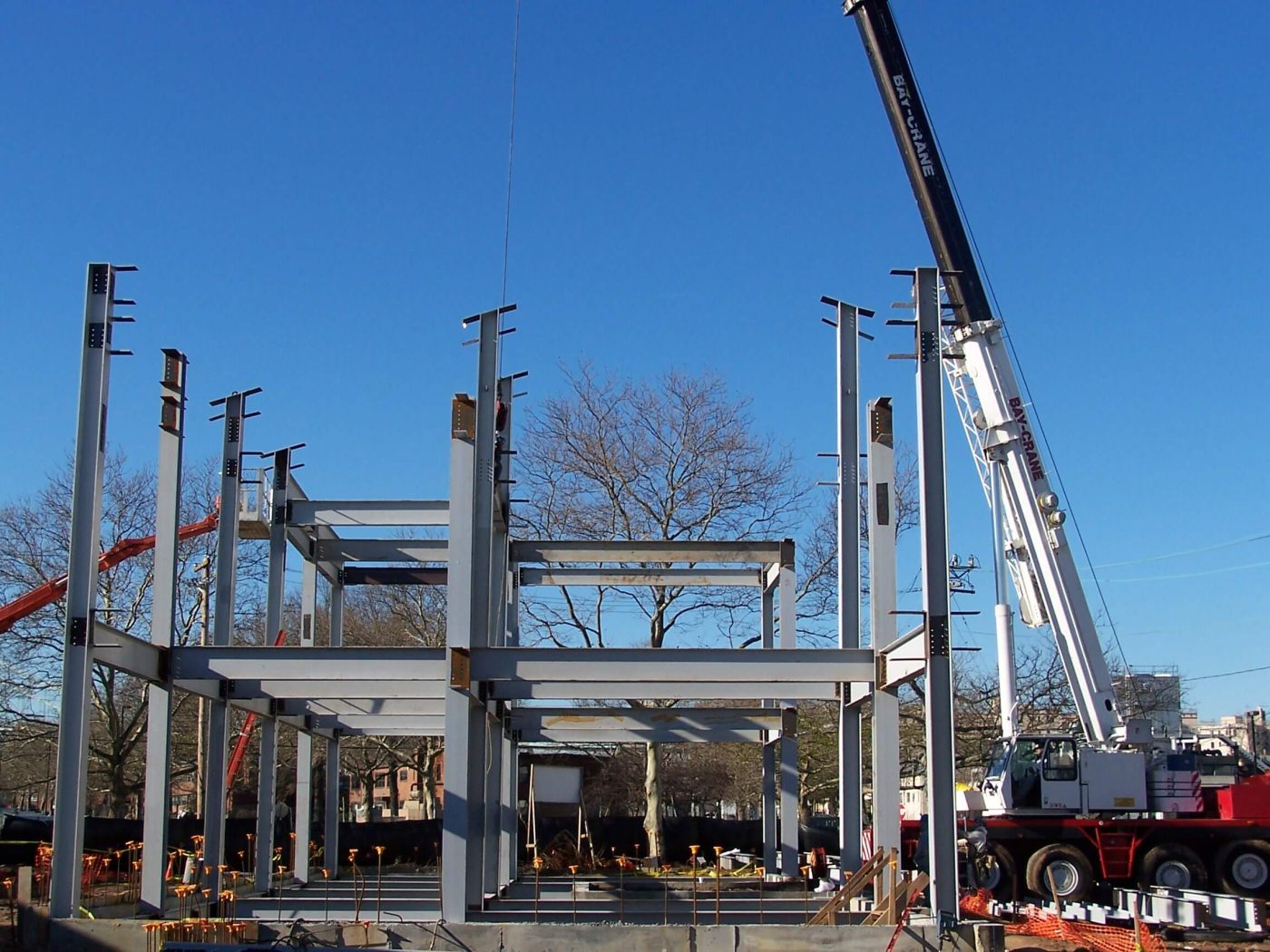 Steel columns with connection clips and the first tier of beams standing against a deep blue sky, with a telescopic crane boom reaching over the frame.