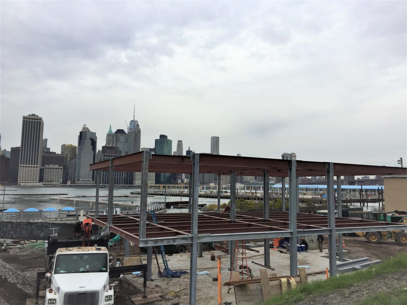 A two-level steel frame of galvanized columns and red primed beams on a waterfront construction site, with a city skyline visible across the water.