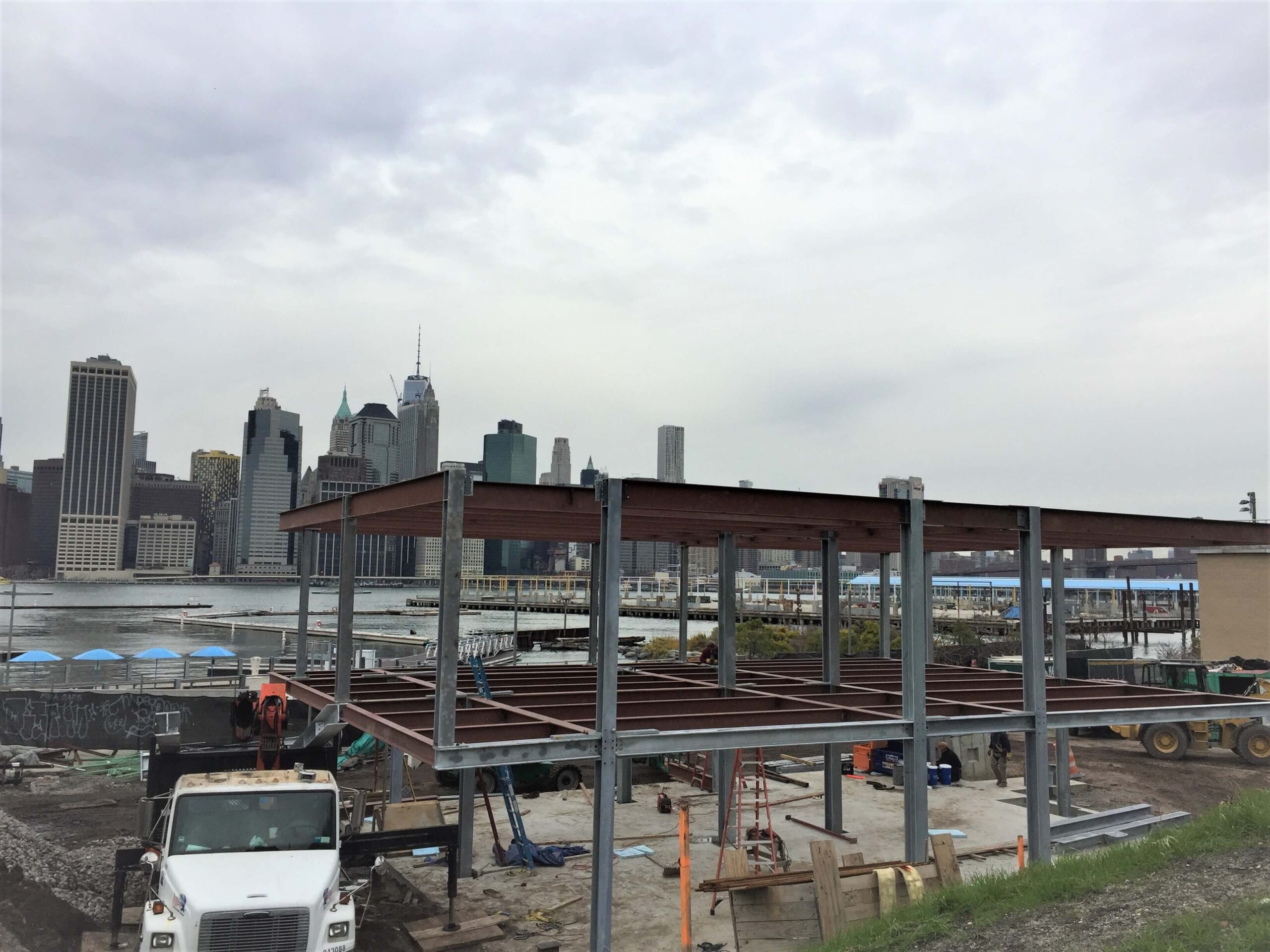 A two-level steel frame of galvanized columns and red primed beams on a waterfront construction site, with a city skyline visible across the water under an overcast sky.