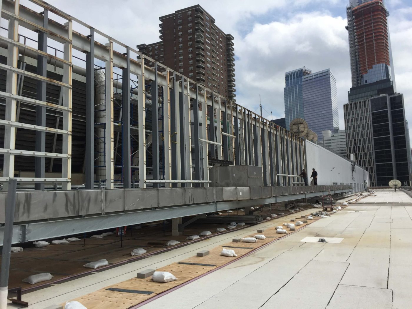 A long line of steel framing for a rooftop mechanical screen wall supported on a plate girder above a flat roof, with high rise towers behind.