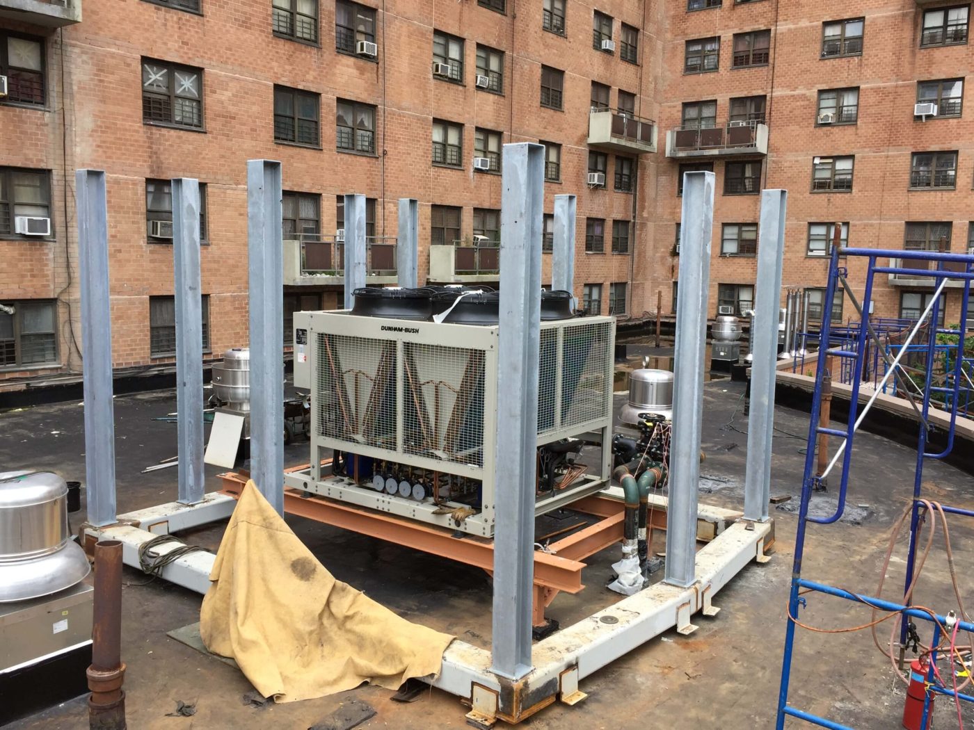 A rooftop air cooled chiller resting on a steel dunnage frame of painted beams with galvanized steel columns standing around it, a brick apartment building behind.