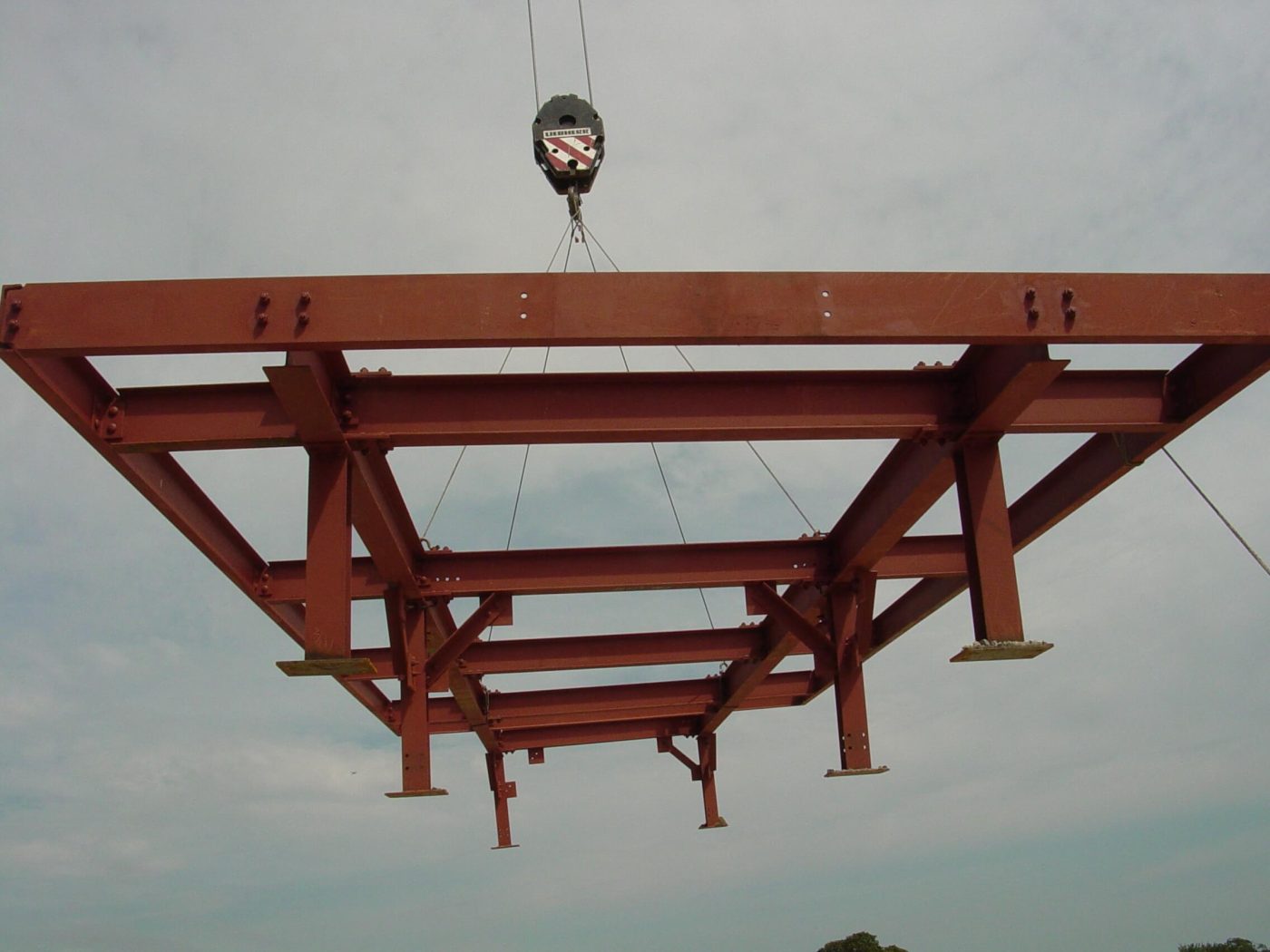 A red primed bolted steel platform frame with short legs and base plates hanging level from a crane block against a pale sky.