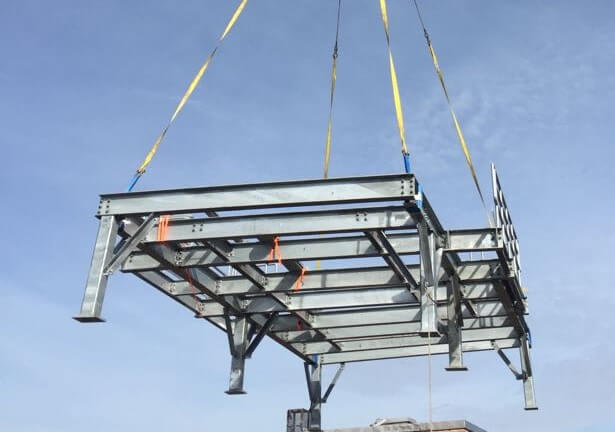 A galvanized steel platform frame slung from four lifting straps on a crane hook against a blue sky.