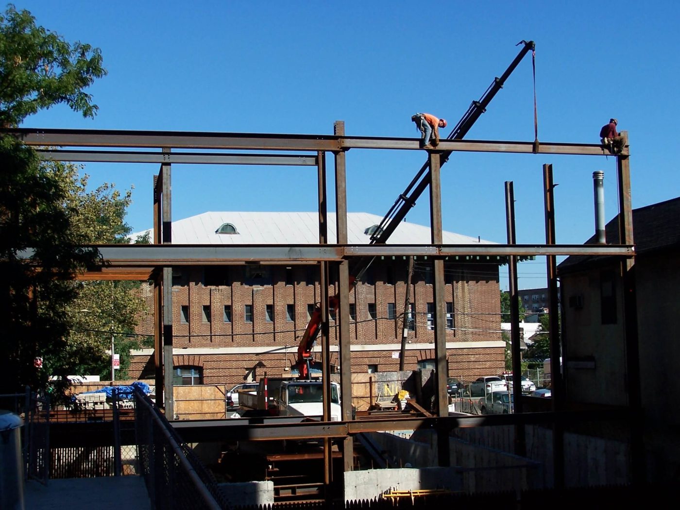 Two ironworkers standing on the top beams of a bare structural steel frame against a deep blue sky while a boom truck holds a beam in place.
