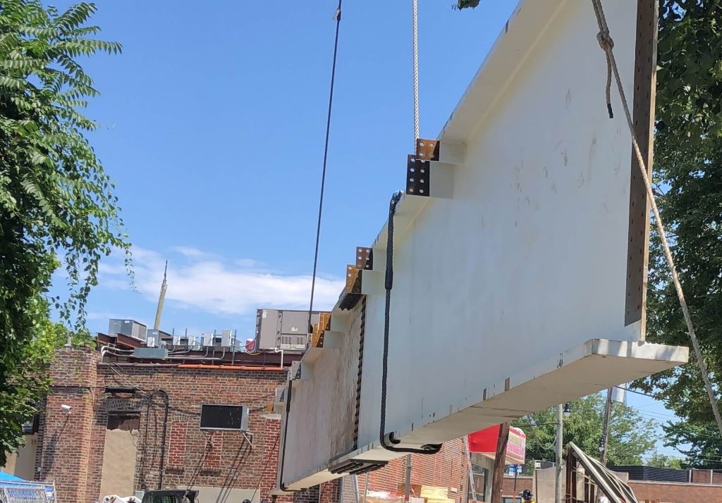 A deep white painted plate girder with welded stiffeners and bolted connection angles hanging from a wire rope sling above a brick storefront on a sunny day.