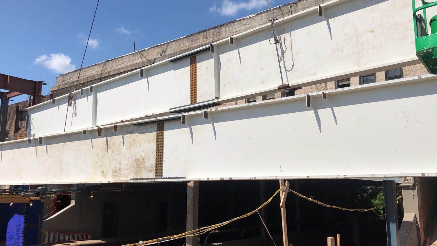 Two long white painted plate girders set side by side and joined with bolted splice plates, spanning above an open storefront under a blue sky.