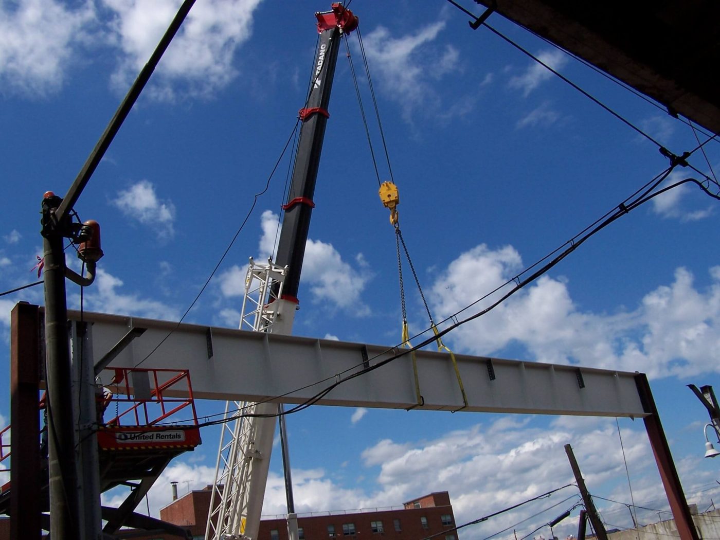 A hydraulic crane lifting a painted wide flange beam on chain slings into a steel frame against a blue sky with scattered clouds, a worker in a boom lift basket at the left.