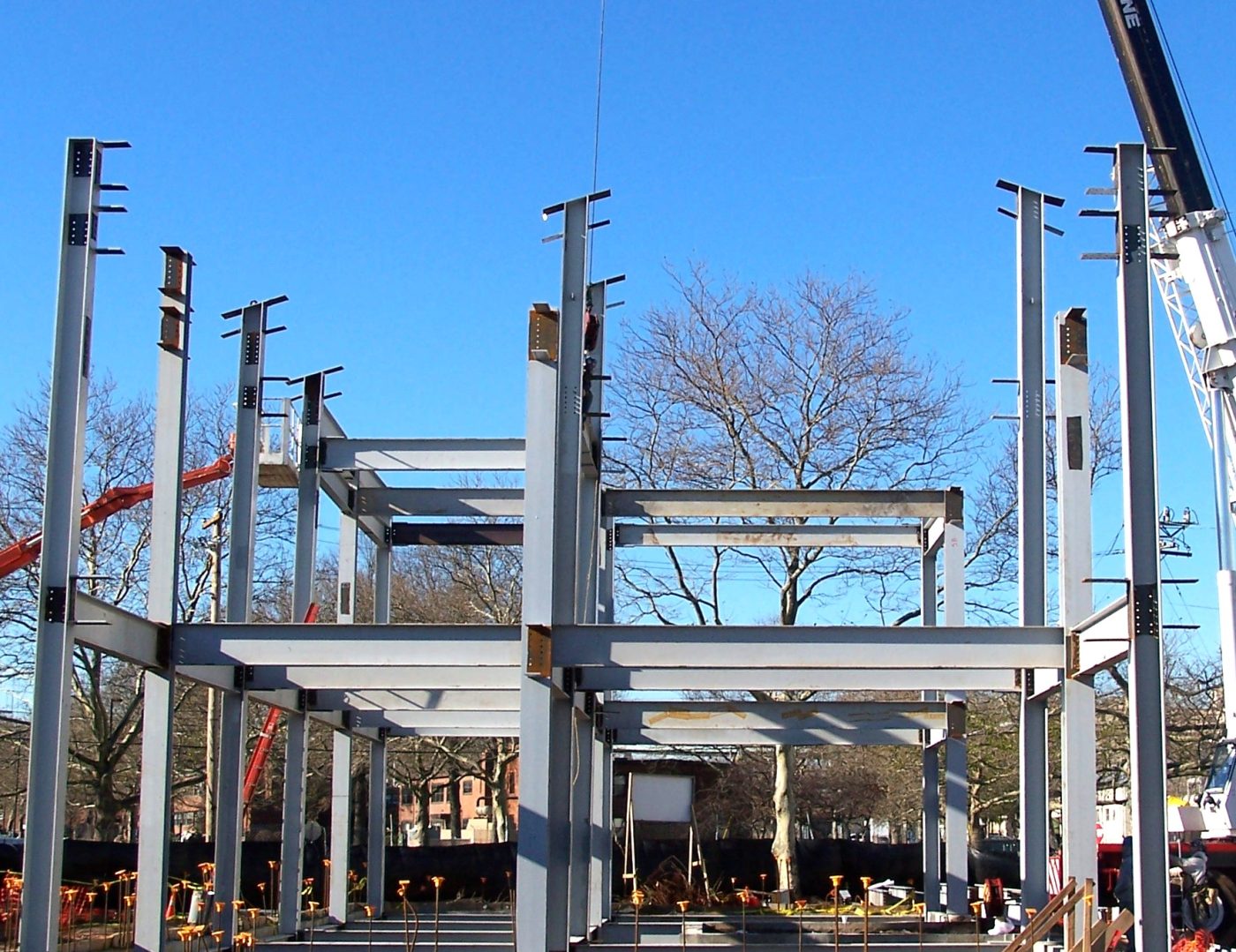 Painted steel columns and beams standing on a concrete slab with anchor bolts in the foreground and a crane at the right.