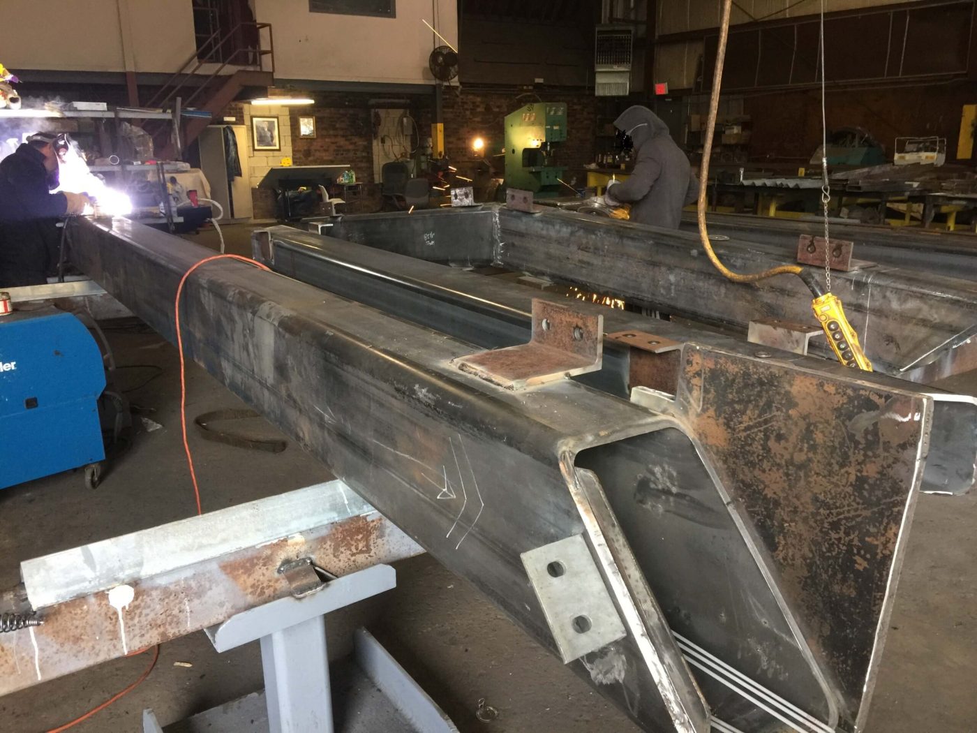 Long fabricated steel box members on stands in a shop, with a welder striking a bright arc at the left and a second worker sending up sparks in the background.